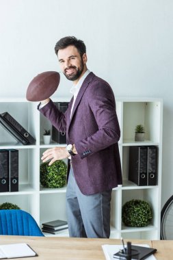 handsome young businessman playing with american football ball at office clipart