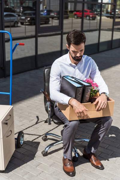 young sad dismissed manager with box of personal stuff sitting on chair outdoors