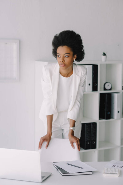 attractive african american businesswoman leaning on chair and looking away in office