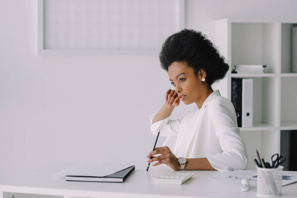 attractive african american businesswoman sitting at table and looking away in office