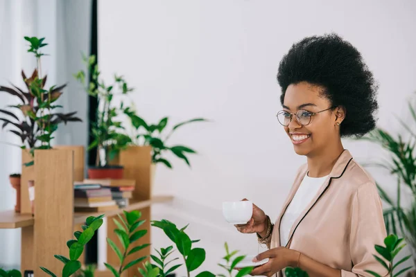 Χαμογελώντας Ελκυστική Αφρικανικός Αμερικανός Επιχειρηματίας Κρατώντας Κούπα Καφέ Στο Γραφείο — Δωρεάν Φωτογραφία
