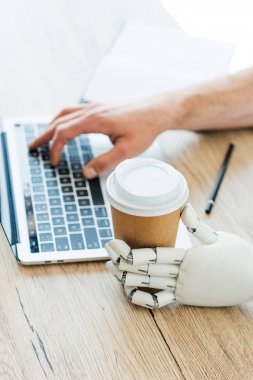 Robotik Kol tutan tek kullanımlık kahve fincanı ve insan eli tahta masada dizüstü bilgisayar kullanarak
