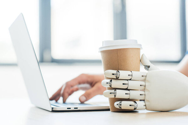close-up view of robotic arm holding disposable coffee cup and human hand using laptop  