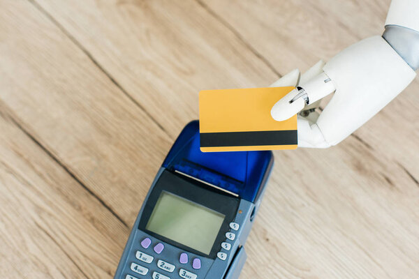 top view of robot holding credit card above payment terminal on wooden table  