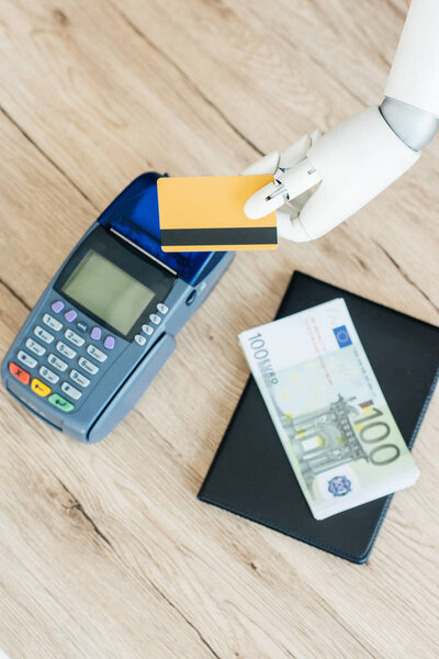 top view of hand of robot holding credit card above payment terminal and money on wooden table 