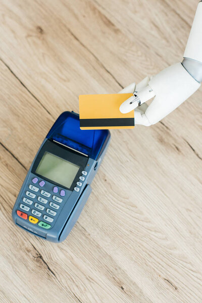 top view of hand of robot holding credit card above payment terminal on wooden table  