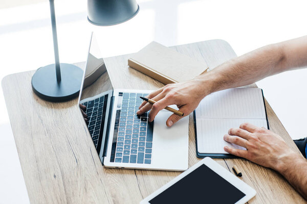 cropped shot of person holding pen, using laptop and taking notes at workplace