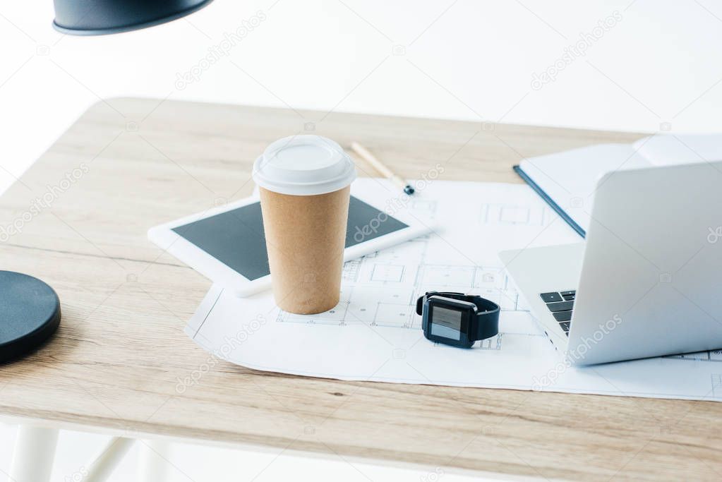 disposable coffee cup, digital tablet, smartwatch, laptop and blueprint on wooden table