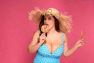 beautiful size plus model in swimsuit holding lollipops and looking at camera isolated on pink clipart