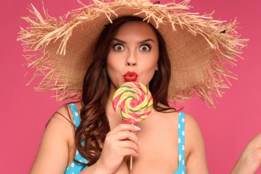 portrait of surprised woman in wicker hat and swimsuit holding lollipop and looking at camera isolated on pink clipart