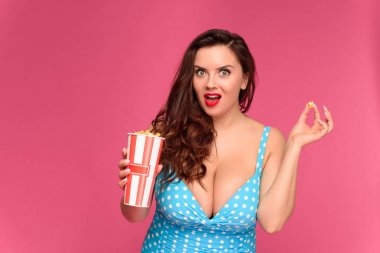 surprised young woman in swimsuit holding box of popcorn and looking at camera clipart