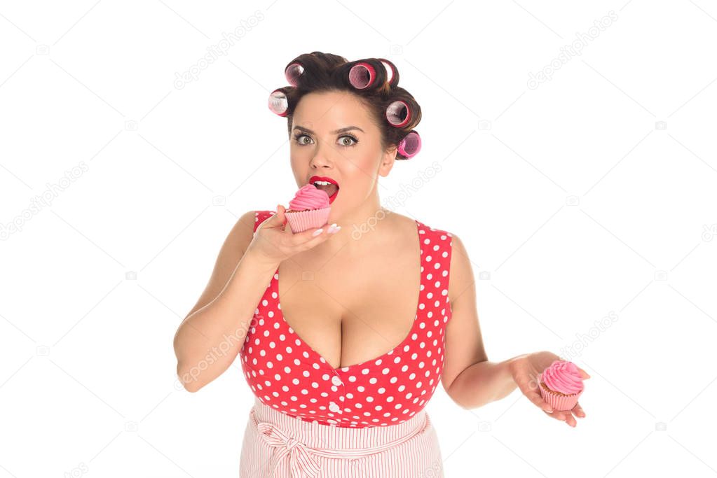emotional plus size woman eating pink cream cupcakes and looking at camera isolated on white