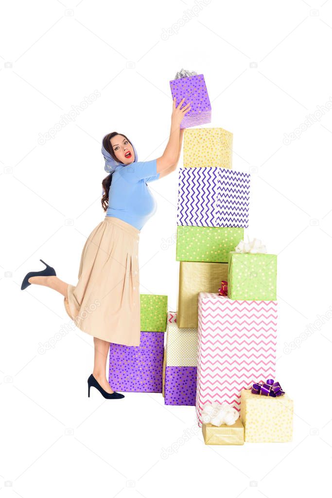 full length view of stylish young woman looking at camera while standing near pile of gift boxes isolated on white
