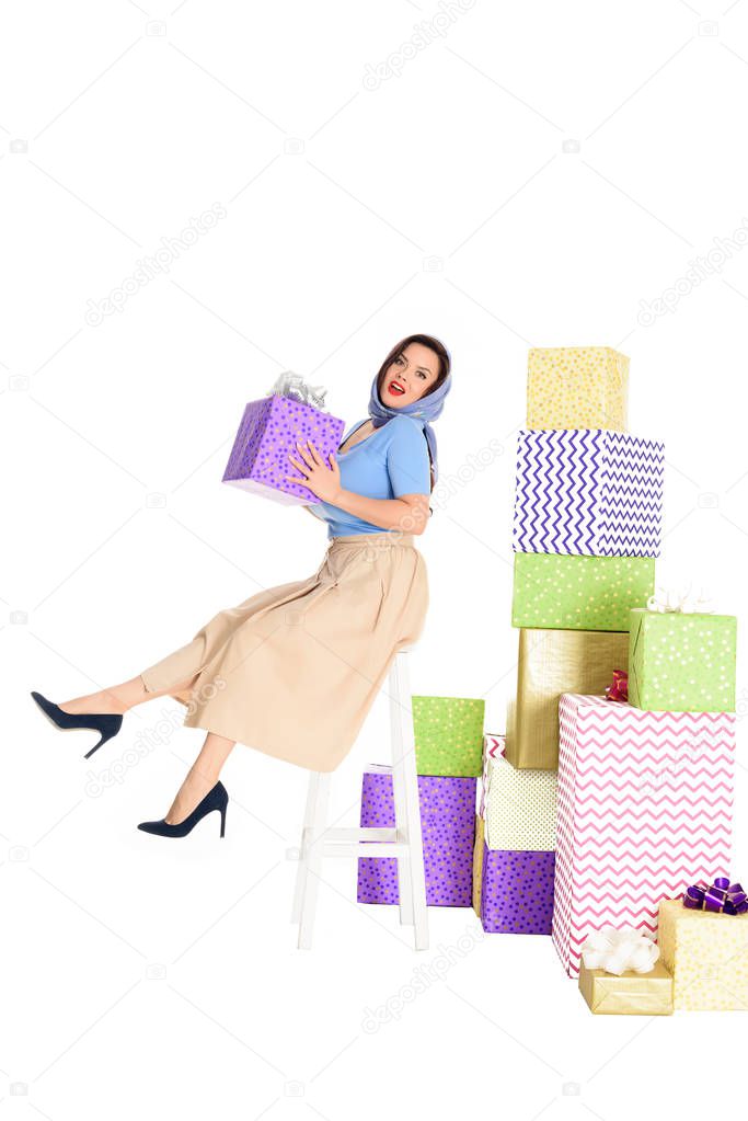 young pin up woman holding gift box and sitting on stool near piles of presents isolated on white  