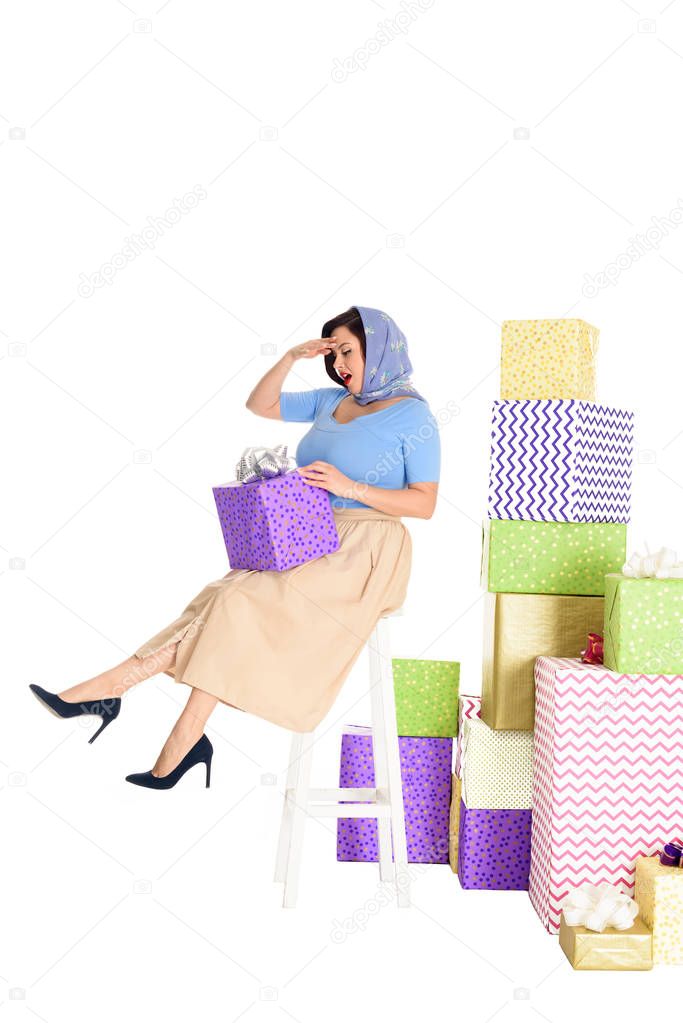 full length view of pin up woman holding gift box and sitting on stool near piles of presents isolated on white 