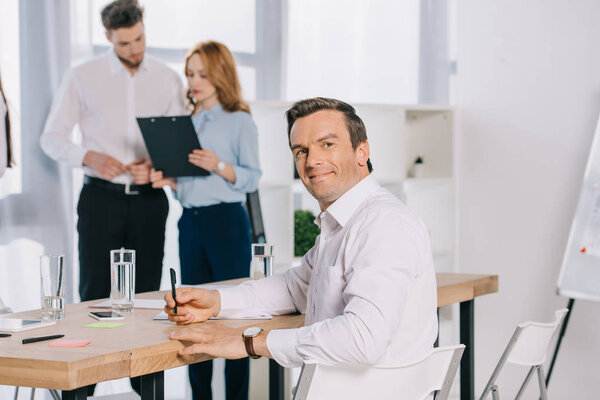 selective focus of smiling businessman at workplace and colleagues discussing work behind in office