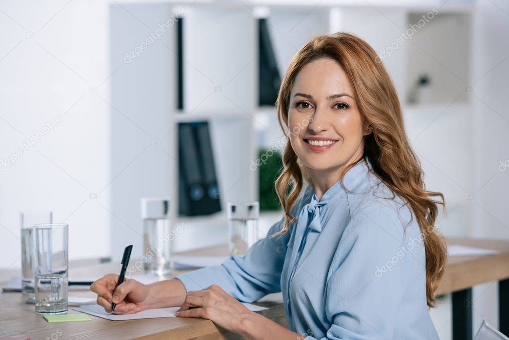 side view of smiling businesswoman looking at camera at workplace with papers in office