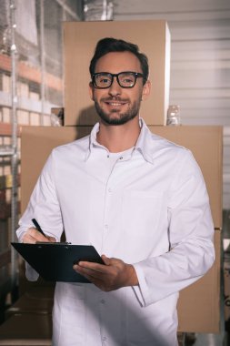 handsome storekeeper in glasses smiling at camera while writing on clipboard clipart