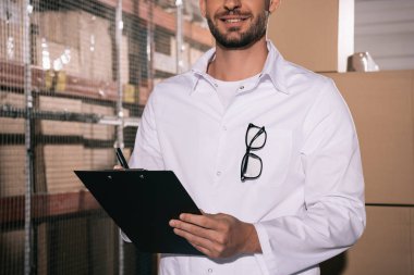 partial view of smiling storekeeper holding clipboard in warehouse clipart