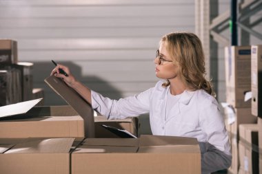 focused storekeeper in white coat inspecting cardboard boxes in warehouse clipart