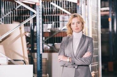 confident businesswoman looking at camera while standing in warehouse with crossed arms clipart