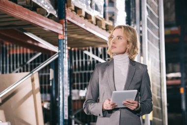 confident businesswoman holding digital tablet while looking away in warehouse clipart