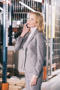 serious businesswoman talking on smartphone and looking away in warehouse clipart