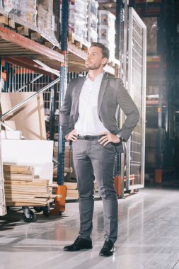 confident businessman looking away while standing in warehouse with hands on hips clipart