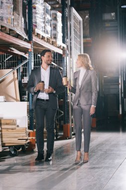 two smiling businesspeople talking while walking in warehouse with coffee to go clipart