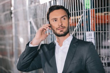confident businessman looking away while talking on smartphone in warehouse clipart
