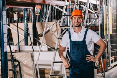 handsome warehouse worker standing with hands on hips and smiling at camera clipart