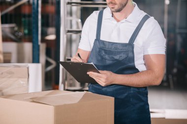 cropped view of warehouse worker writing on clipboard while standing near cardboard box clipart