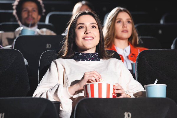 избирательный фокус улыбающейся женщины, держащей попкорн и смотрящей кино в кино
