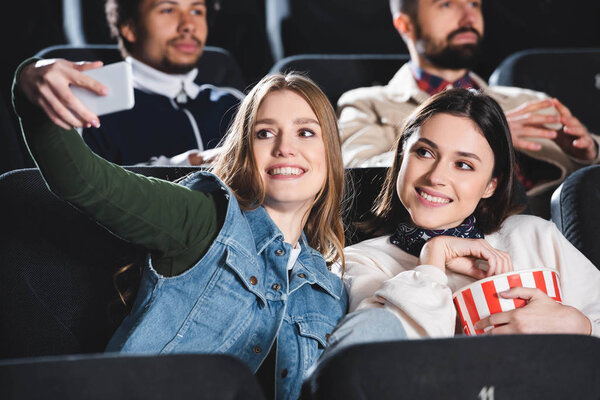 selective focus of smiling friends with popcorn taking selfie in cinema 