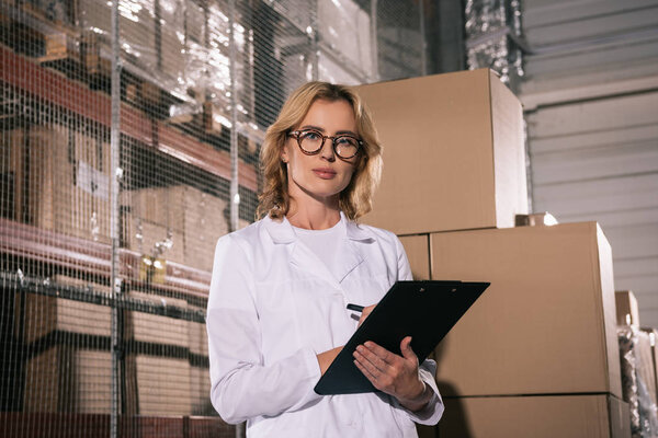 serious storekeeper looking at camera while writing on clipboard in warehouse
