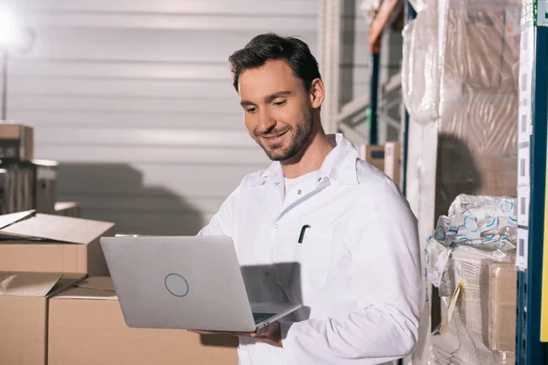 Smiling Storekeeper Using Laptop While Standing Cardboard Boxes Storehouse — Stock Photo, Image