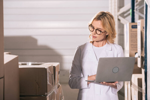 concentrated storekeeper using laptop while looking at cardboard boxes in warehouse
