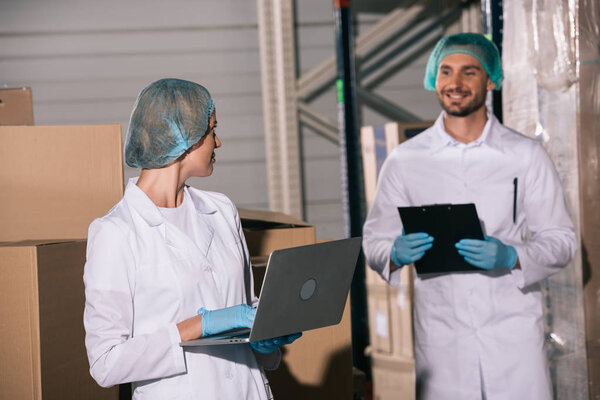 smiling storekeeper holding clipboard and looking at colleague using laptop in warehouse