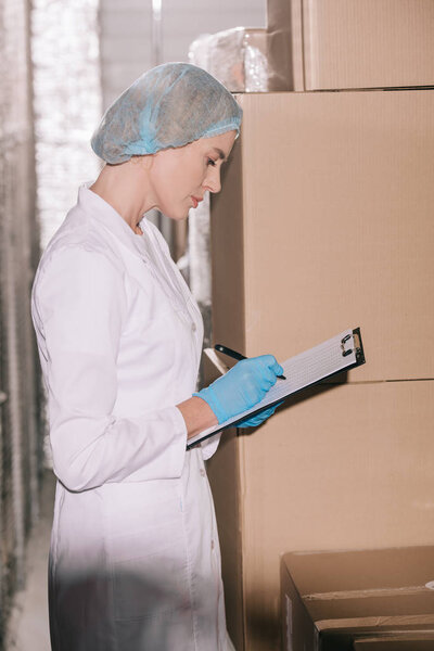 focused storekeeper in hairnet writing on clipboard while standing near carton boxes in warehouse
