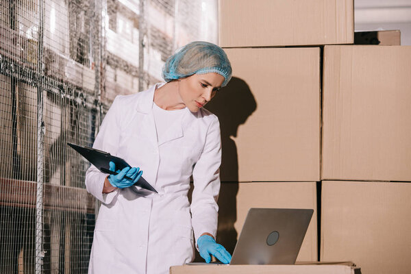 concentrated storekeeper in white coat and hairnet using laptop and holding clipboard 