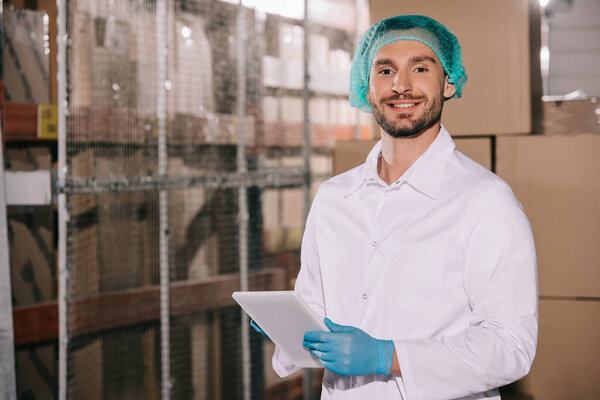 cheerful storekeeper in white coat and hairnet holding digital tablet and looking at camera