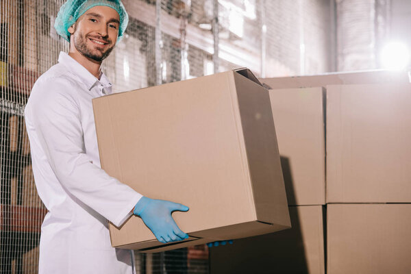 cheerful storekeeper smiling at camera while holding cardboard box in warehouse