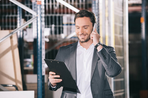 smiling businessman talking on smartphone and looking on clipboard in warehouse