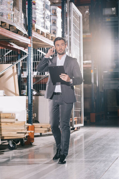serious businessman talking on smartphone and holding clipboard in warehouse
