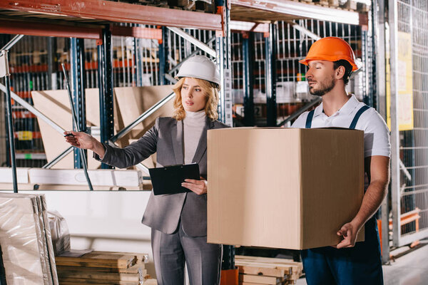 businesswoman in helmet pointing with hand while standing near loader holding cardboard box