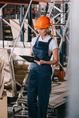 selective focus of workwoman in overalls and helmet standing near construction materials and writing on clipboard clipart