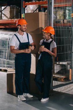 Gülümseyen depo çalışanları birbirlerine bakıp kahve içiyorlar.