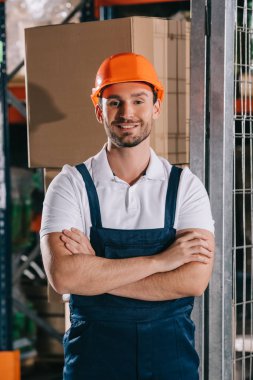 handsome warehouse worker smiling at camera while standing with crossed arms clipart