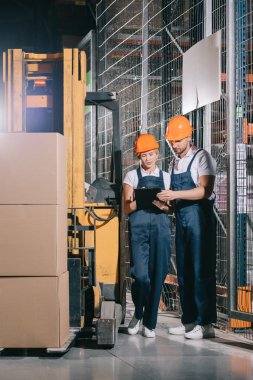 two warehouse workers looking at clipboard while standing near forklift loader clipart
