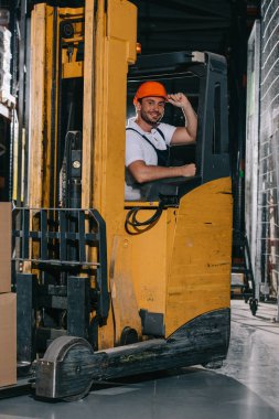 smiling warehouse worker touching helmet and looking at camera while operating forklift loader clipart